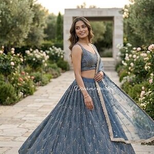May include: A slate blue lehenga choli set featuring a sleeveless blouse and a voluminous floor-length skirt, both adorned with intricate silver geometric embroidery. A matching sheer dupatta with a gold-toned border is draped over one shoulder.