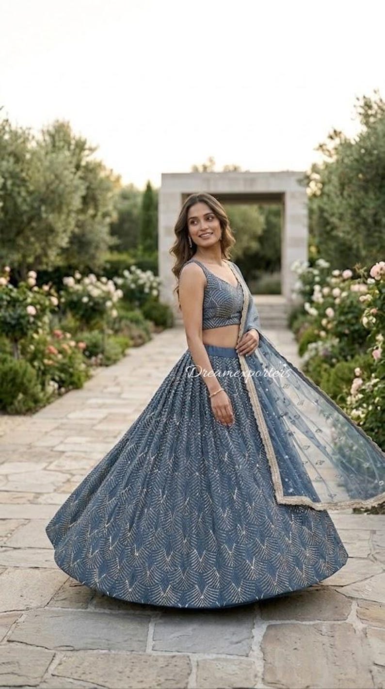 May include: A slate blue lehenga choli set featuring a sleeveless blouse and a voluminous floor-length skirt, both adorned with intricate silver geometric embroidery. A matching sheer dupatta with a gold-toned border is draped over one shoulder.