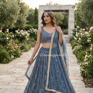 May include: A slate blue lehenga choli set featuring a sleeveless blouse and a floor-length skirt, both adorned with intricate silver geometric embroidery. A matching sheer dupatta with a gold-toned border is draped over the shoulder. Text reads Dreamexporters.