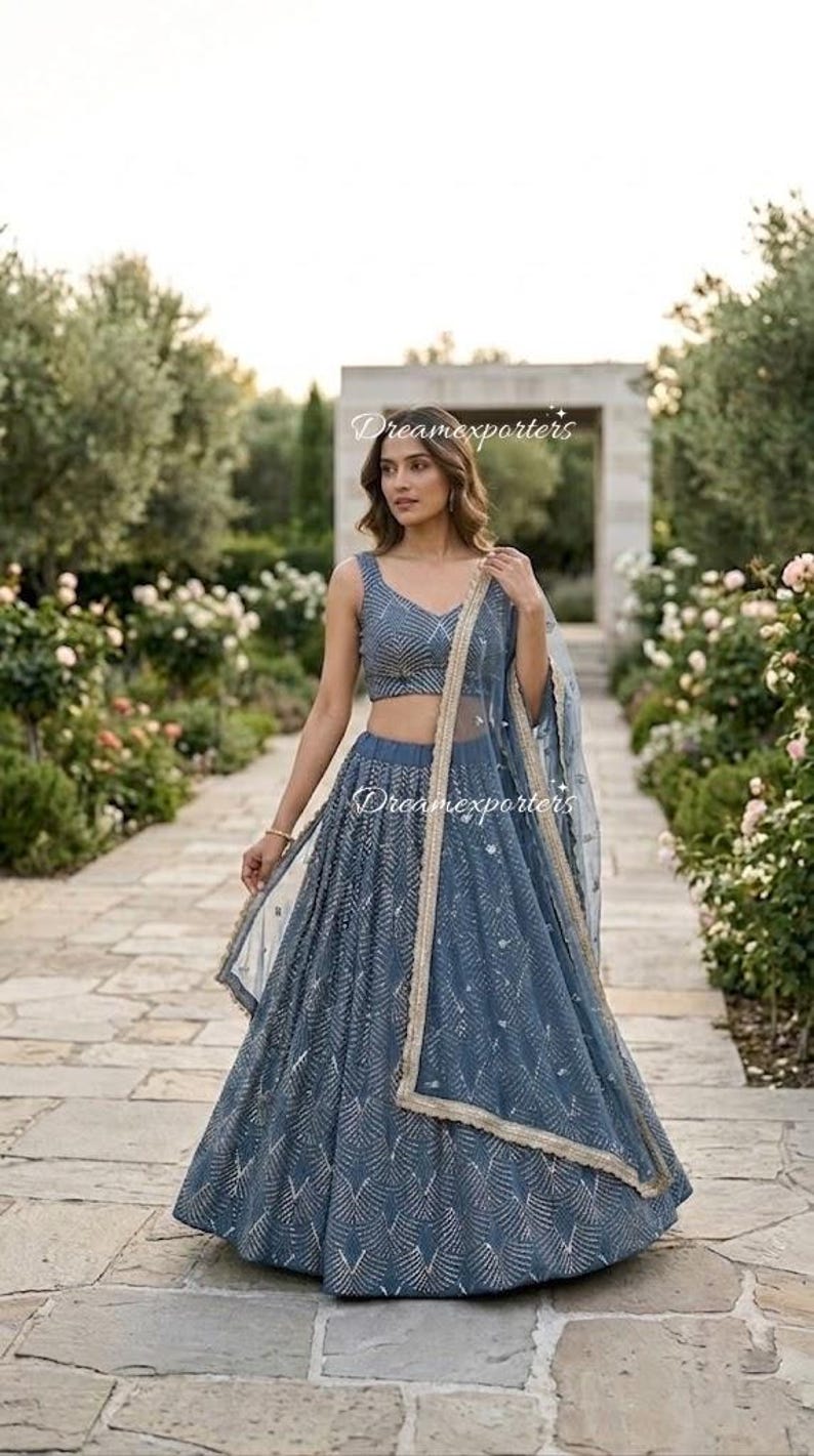 May include: A slate blue lehenga choli set featuring a sleeveless blouse and a floor-length skirt, both adorned with intricate silver geometric embroidery. A matching sheer dupatta with a gold-toned border is draped over the shoulder. Text reads Dreamexporters.
