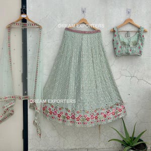 Op de afbeelding: Een lichtgroene geborduurde rok met een bijpassend lichtgroen mouwloos blouse en een doorschijnende lichtgroene dupatta. De rok heeft een bloemenpatroon met roze en rode bloemen. De tekst "DREAM EXPORTERS" is zichtbaar op de afbeelding.