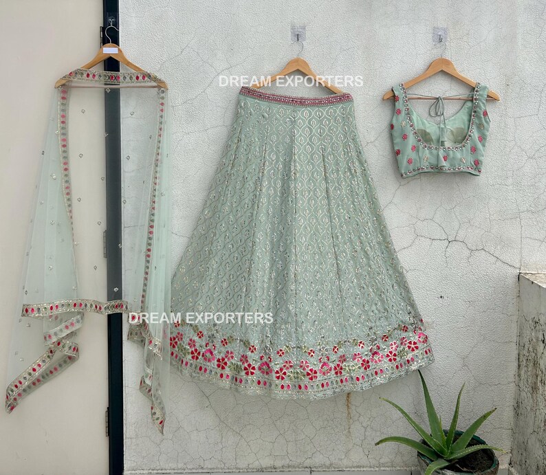 Op de afbeelding: Een lichtgroene geborduurde rok met een bijpassend lichtgroen mouwloos blouse en een doorschijnende lichtgroene dupatta. De rok heeft een bloemenpatroon met roze en rode bloemen. De tekst "DREAM EXPORTERS" is zichtbaar op de afbeelding.