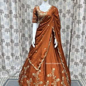 Könnte beinhalten: Ein rostfarbenes formelles Kleid mit aufwendiger goldener Stickerei und Verzierungen. Das Kleid hat einen passenden Schal und ein detailliertes Oberteil. Der Text "Dream Exporters" ist auf dem Rock zu sehen.