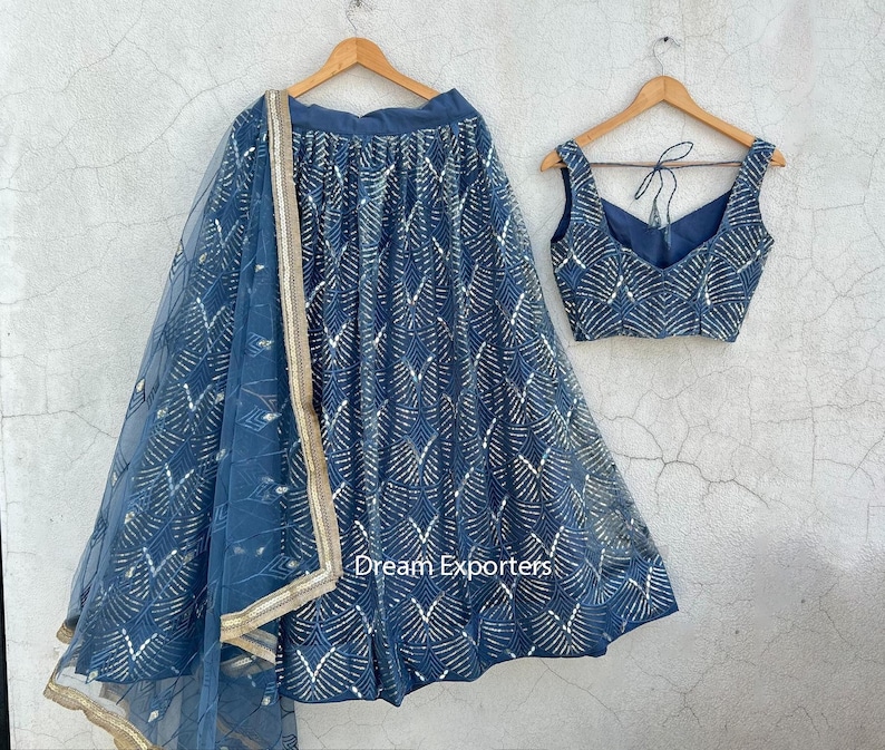 Puede incluir: Un conjunto de dos piezas azul, compuesto por una falda y un top, ambos adornados con un patr&oacute;n geom&eacute;trico dorado repetido. La falda tiene un dise&ntilde;o fluido, y el top presenta un cuello en V. El conjunto incluye una bufanda transparente a juego con ribete dorado.