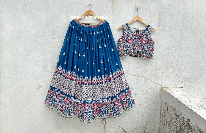 May include: A two-piece ensemble featuring a blue skirt and a matching top. The skirt is pleated and adorned with a detailed floral pattern in shades of red, white, and green. The top has a similar floral design.