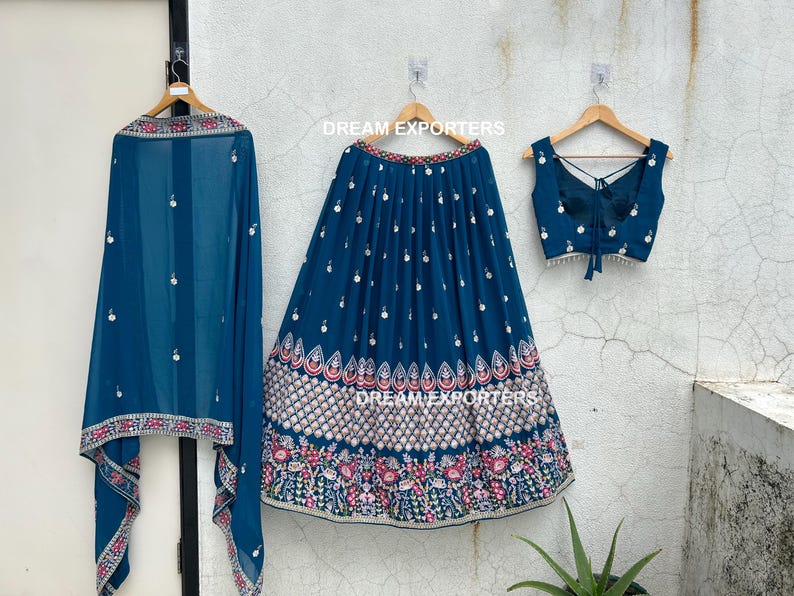 May include: A three-piece teal ensemble featuring a skirt, top, and scarf. The skirt and scarf have a striped pattern with white embellishments. The top is sleeveless with a tie-back design. The skirt and scarf have a floral border.