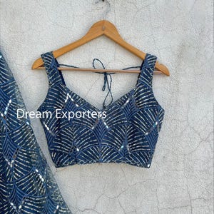 Puede incluir: Un top azul marino con cuello en V y tirantes anchos, adornado con lentejuelas plateadas en forma de abanico. El texto "Dream Exporters" es visible. El top est&aacute; colgado de una percha de madera.