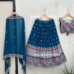 May include: A three-piece teal-colored Indian outfit, including a skirt, top, and scarf, all adorned with intricate embroidery and embellishments. The skirt and top feature floral patterns in pink, white, and gold. The scarf is sheer and has a matching border.