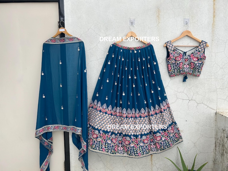 May include: A three-piece teal-colored Indian outfit, including a skirt, top, and scarf, all adorned with intricate embroidery and embellishments. The skirt and top feature floral patterns in pink, white, and gold. The scarf is sheer and has a matching border.