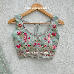 Op de afbeelding: Een lichtgroen, mouwloos blouse met een ronde hals en een strik aan de achterkant. De blouse is geborduurd met roze en groene bloemenmotieven en versierd met pailletten. De blouse hangt aan een houten hanger met de tekst "DREAM EXPORTERS" erop gedrukt.