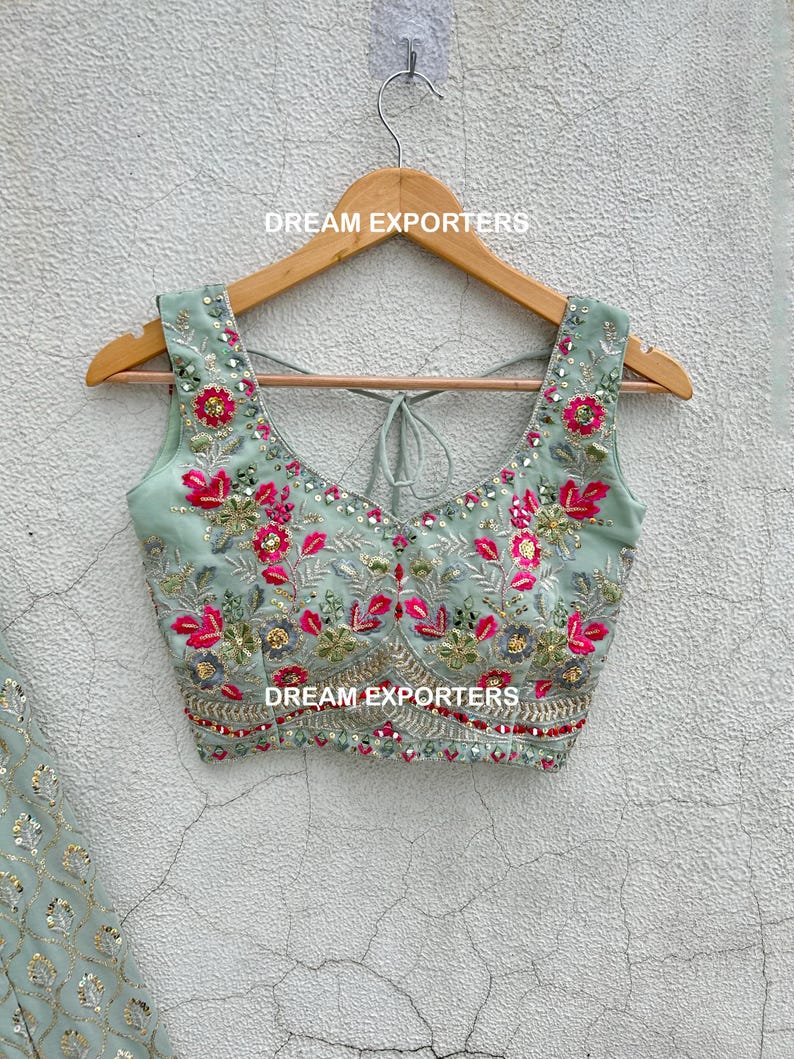Op de afbeelding: Een lichtgroen, mouwloos blouse met een ronde hals en een strik aan de achterkant. De blouse is geborduurd met roze en groene bloemenmotieven en versierd met pailletten. De blouse hangt aan een houten hanger met de tekst "DREAM EXPORTERS" erop gedrukt.