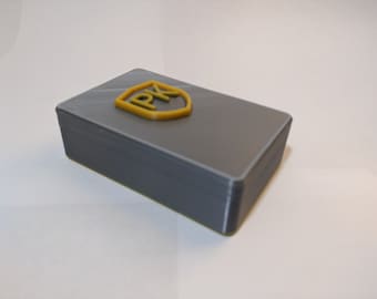 Puede incluir: Una caja rectangular gris con un logotipo de escudo amarillo en relieve que dice "PK".