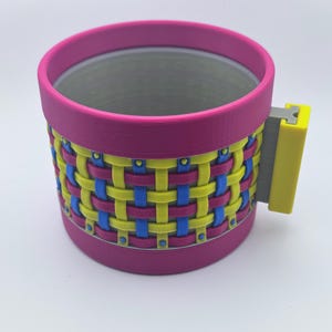 May include: A pink and yellow woven cup holder with a blue and yellow design. The holder has a gray and yellow handle.
