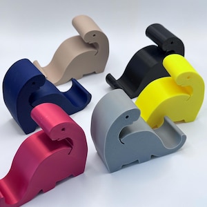 Op de afbeelding: Een verzameling kleurrijke telefoonstandaards in de vorm van een dinosaurus in verschillende kleuren, waaronder roze, blauw, beige, zwart, geel en grijs. Elke standaard is ontworpen om een telefoon vast te houden en heeft een gebogen vorm.