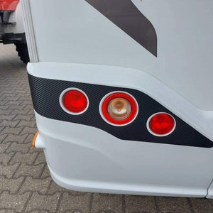 Puede incluir: Autocaravana blanca con un acabado negro de fibra de carbono en el parachoques trasero. El parachoques tiene tres luces traseras rojas y una luz de se&ntilde;alizaci&oacute;n &aacute;mbar.