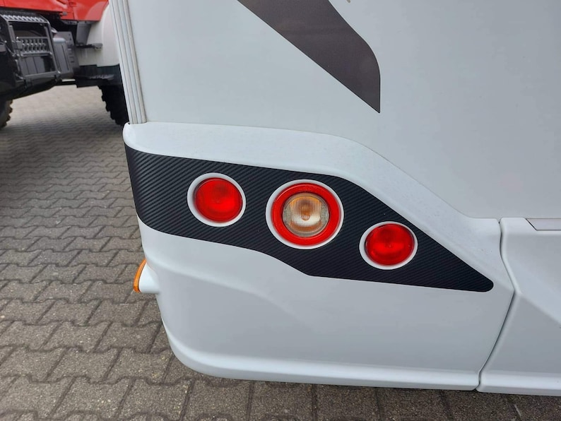 Puede incluir: Autocaravana blanca con un acabado negro de fibra de carbono en el parachoques trasero. El parachoques tiene tres luces traseras rojas y una luz de se&ntilde;alizaci&oacute;n &aacute;mbar.