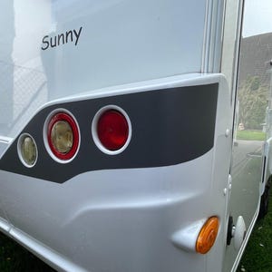 Puede incluir: Una autocaravana blanca con una franja gris y la palabra "Sunny" en el lateral. La autocaravana tiene tres luces traseras rojas y una luz de se&ntilde;alizaci&oacute;n amarilla.