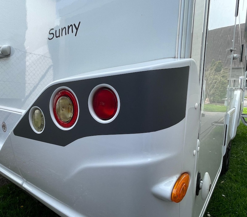 Puede incluir: Una autocaravana blanca con una franja gris y la palabra "Sunny" en el lateral. La autocaravana tiene tres luces traseras rojas y una luz de se&ntilde;alizaci&oacute;n amarilla.