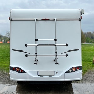 Puede incluir: Una autocaravana blanca con un portabicicletas plateado en la parte trasera. El portabicicletas tiene dos barras horizontales y una escalera.