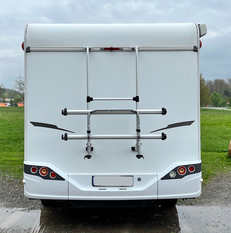 Puede incluir: Una autocaravana blanca con un portabicicletas plateado en la parte trasera. El portabicicletas tiene dos barras horizontales y una escalera.