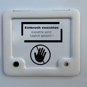 Peut inclure: Porte blanche avec une pancarte noire et blanche indiquant "Einbruch zwecklos Kassette wird täglich geleert!" avec une main noire barrée d'une ligne diagonale.