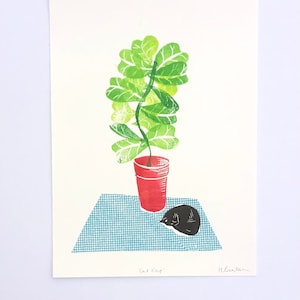Puede incluir: Un grabado en linóleo que representa una planta verde en una maceta roja, sobre una superficie a cuadros azules. Un gato negro durmiendo está al lado de la maceta. La impresión se titula "Cat Nap" y está firmada por el artista.