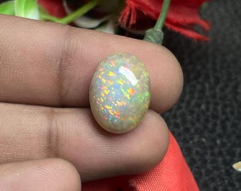 Può includere: Una gemma di opale di forma ovale con una base bianco latte. L'opale mostra un vivace gioco di colori, con lampi di arancione, giallo e verde. La gemma è tenuta tra le dita, con fiori rossi sullo sfondo.