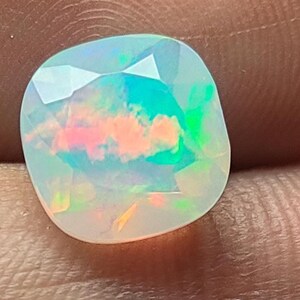 May include: A faceted, oval-shaped opal gemstone. The opal displays a play of color with iridescent hues of pink, green, and blue. The gemstone is held in a hand, showcasing its polished surface and internal patterns.