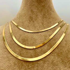Puede incluir: Un collar de cadenas de espiga en tono dorado superpuestas, compuesto por tres hilos separados de longitudes variables. Cada cadena presenta un diseño de metal plano, flexible y pulido que refleja la luz sobre su superficie lisa.