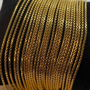 May include: A collection of multiple gold-toned box chains arranged side by side. Each chain features a series of small, square, interlocking links that create a smooth, uniform, and flexible metallic strand with a reflective, polished finish.