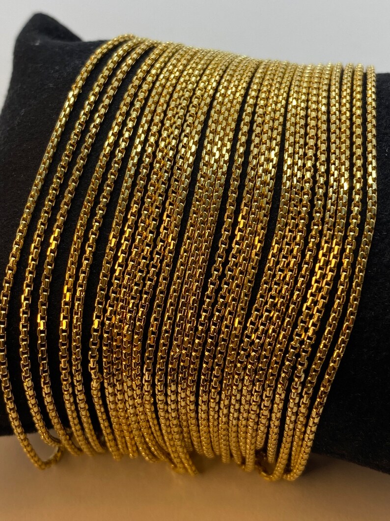May include: A collection of multiple gold-toned box chains arranged side by side. Each chain features a series of small, square, interlocking links that create a smooth, uniform, and flexible metallic strand with a reflective, polished finish.