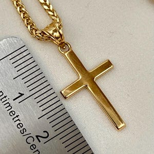 May include: A gold cross pendant on a matching chain. The cross has a simple, classic design with a polished finish. The chain is a wheat-link style. A ruler is in the image, showing centimetres.