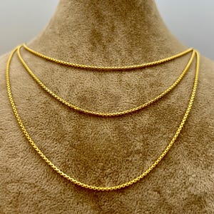 May include: A layered gold-tone necklace featuring three distinct chains with a rounded, textured link design. The chains are arranged in a cascading style, each displaying a consistent metallic yellow finish and a uniform, interconnected circular pattern.