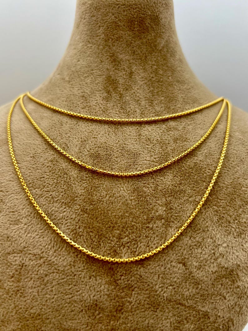 May include: A layered gold-tone necklace featuring three distinct chains with a rounded, textured link design. The chains are arranged in a cascading style, each displaying a consistent metallic yellow finish and a uniform, interconnected circular pattern.