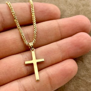 May include: A gold cross pendant on a braided chain. The cross has a simple, rectangular design. The necklace is shown against a neutral background. The chain and pendant appear to be made of gold.