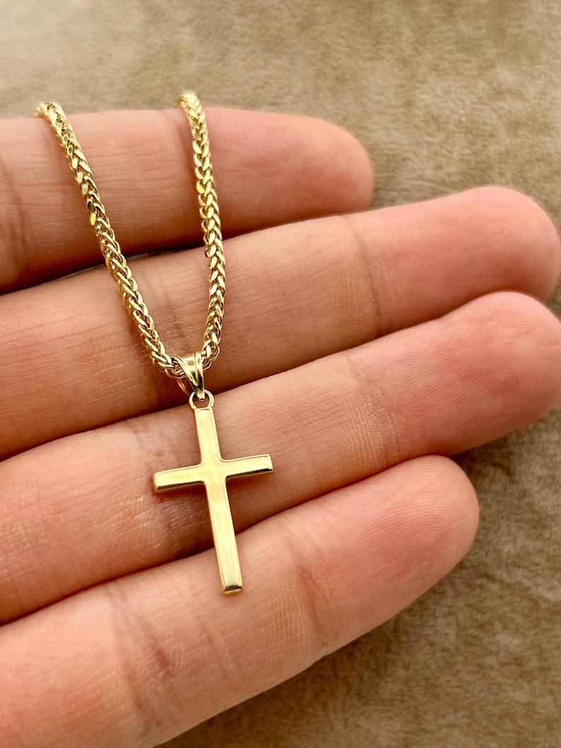 May include: A gold cross pendant on a braided chain. The cross has a simple, rectangular design. The necklace is shown against a neutral background. The chain and pendant appear to be made of gold.