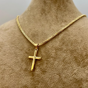 May include: A gold cross pendant necklace. The cross is a simple design, suspended from a matching gold chain. The chain has a woven texture. The necklace is displayed on a neutral-toned mannequin.