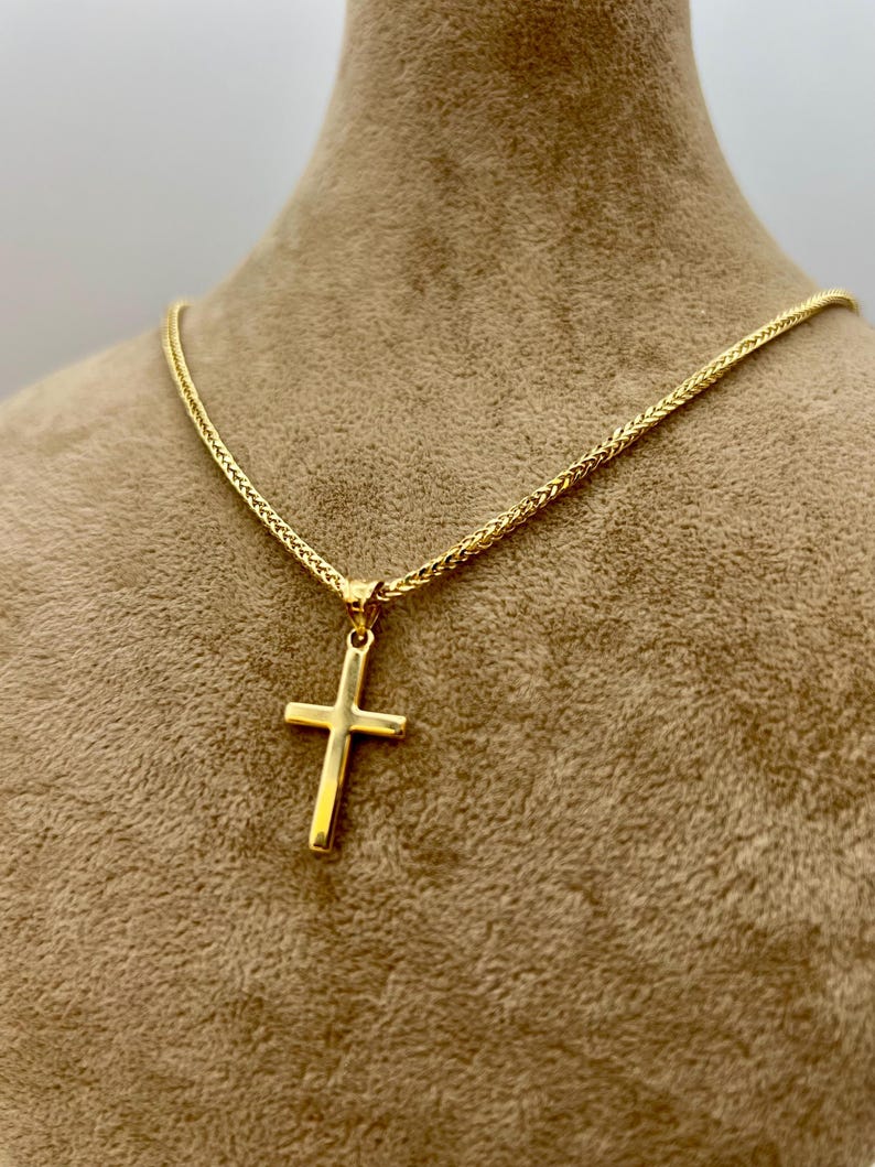May include: A gold cross pendant necklace. The cross is a simple design, suspended from a matching gold chain. The chain has a woven texture. The necklace is displayed on a neutral-toned mannequin.