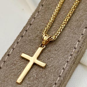 May include: A gold cross pendant necklace. The cross has a simple, classic design with a polished finish. The chain is a woven style, also in gold. The necklace rests on a textured, tan surface.