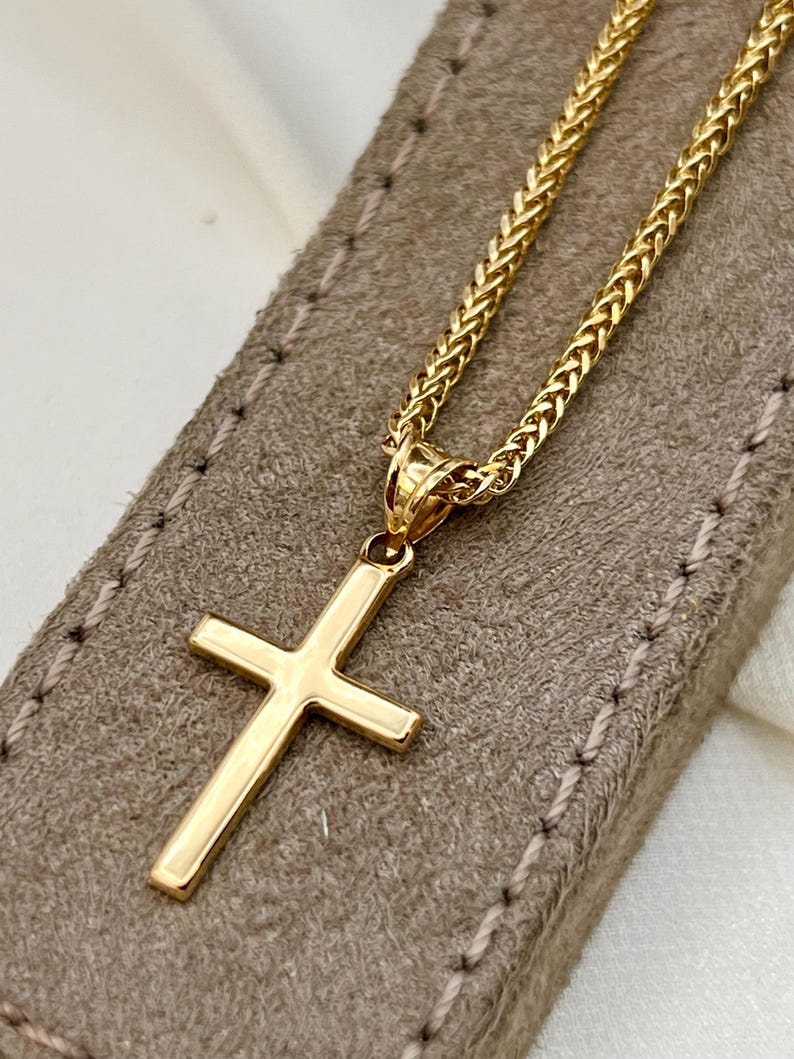 May include: A gold cross pendant necklace. The cross has a simple, classic design with a polished finish. The chain is a woven style, also in gold. The necklace rests on a textured, tan surface.