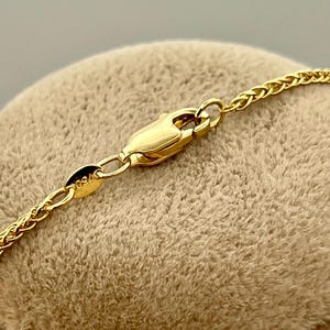 May include: Close-up of a gold-coloured necklace with a wheat chain design. The necklace features a lobster clasp closure. The clasp and chain are made of a shiny, yellow metal. The necklace is resting on a tan, textured surface.