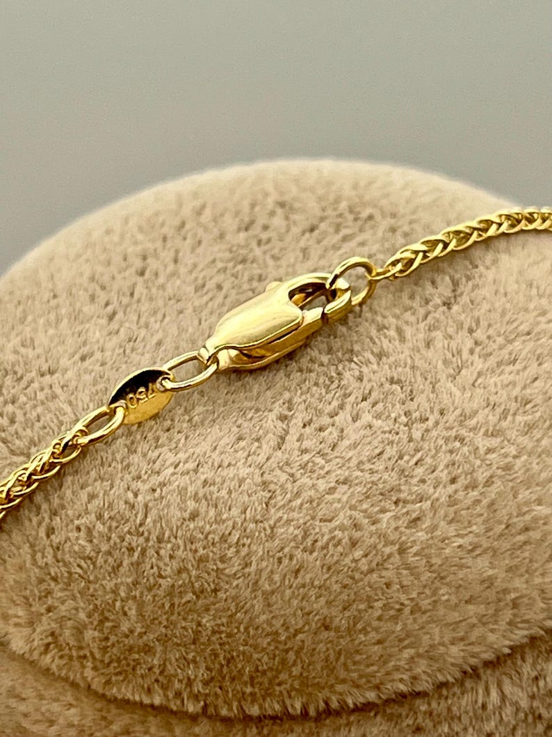 May include: Close-up of a gold-coloured necklace with a wheat chain design. The necklace features a lobster clasp closure. The clasp and chain are made of a shiny, yellow metal. The necklace is resting on a tan, textured surface.