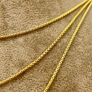 May include: A bright yellow gold chain necklace featuring a box link design. The chain is composed of small, square-shaped metallic links connected in a repeating pattern, creating a smooth, flexible, and continuous strand with a polished finish.