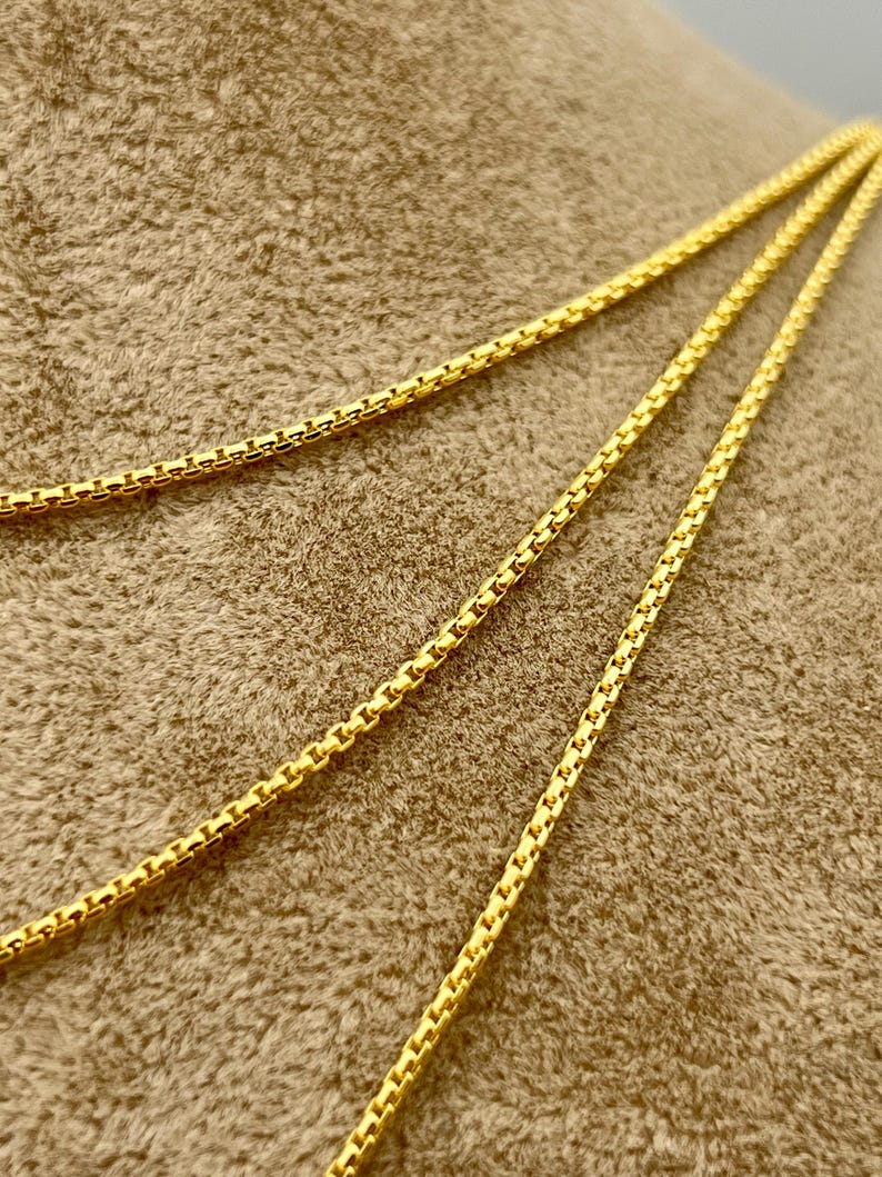May include: A bright yellow gold chain necklace featuring a box link design. The chain is composed of small, square-shaped metallic links connected in a repeating pattern, creating a smooth, flexible, and continuous strand with a polished finish.