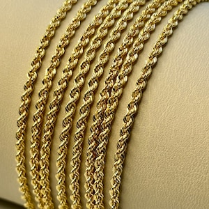 May include: A close-up of a stack of gold rope chains. The chains are arranged in a neat row, with the ends of each chain overlapping the next.