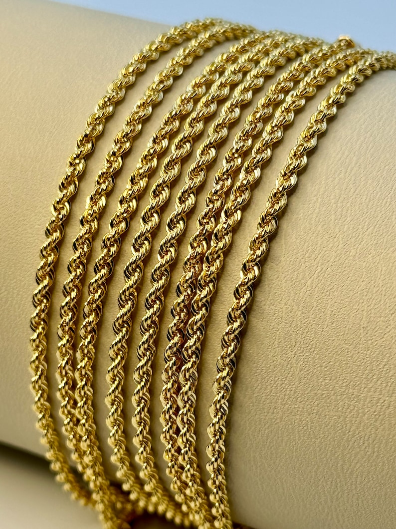 May include: A close-up of a stack of gold rope chains. The chains are arranged in a neat row, with the ends of each chain overlapping the next.