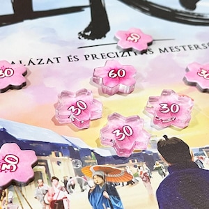 Könnte beinhalten: Ein Brettspiel mit rosa blütenförmigen Spielsteinen, von denen einige die Zahl 30 und einer die 60 anzeigen. Das Spielbrett zeigt eine Illustration im japanischen Stil mit Text in einer Fremdsprache. Die Spielsteine sind verstreut.