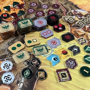 May include: A selection of board game components, such as hexagonal, square, and circular tokens, displayed on a map-style game board. The pieces are in various colours, including green, red, and beige, with symbols and numbers.