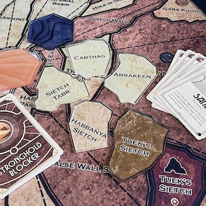 Può includere: Un'impostazione di gioco da tavolo con una mappa, carte e pedine. La mappa è marrone e beige con aree etichettate. Le carte recitano "SAND" e "STRONGHOLD BLOCKER". Le pedine sono blu, arancioni e beige. Il gioco è ambientato su una superficie blu scuro.