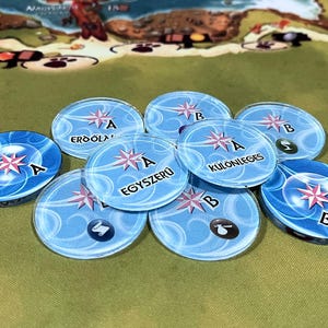 May include: A close-up of eight blue and white circular game pieces with pink compass designs. The pieces have the letters "A" and "B" and the words "EGYszeru", "ERDOOLA", "KULONLEGES" printed on them.